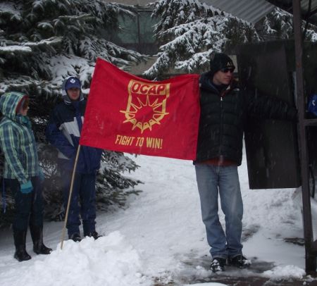 An Ontario Coalition Against Poverty flag and people listening to speeches during the rally portion of the Solidarity Against Austerity event in Memorial Park. (Photo by Scott Neigh)